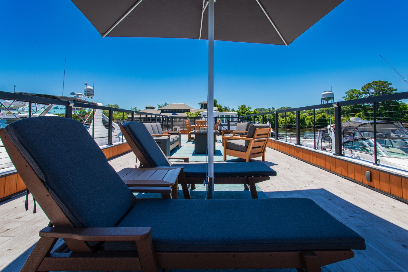 The full rooftop deck looking toward the fire pit and lounge seating, with chaise lounges, a shade umbrella, and the marina visible on both sides under a clear Carolina sky.