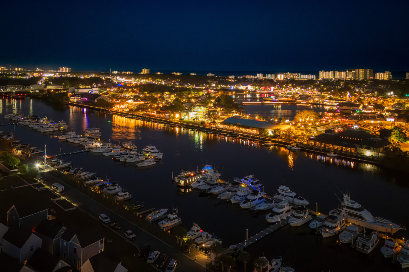 Nighttime aerial of Barefoot Landing Marina and the full waterfront district lit up along the Intracoastal, with Palmera glowing at the dock and the Grand Strand coastline stretching to the horizon.