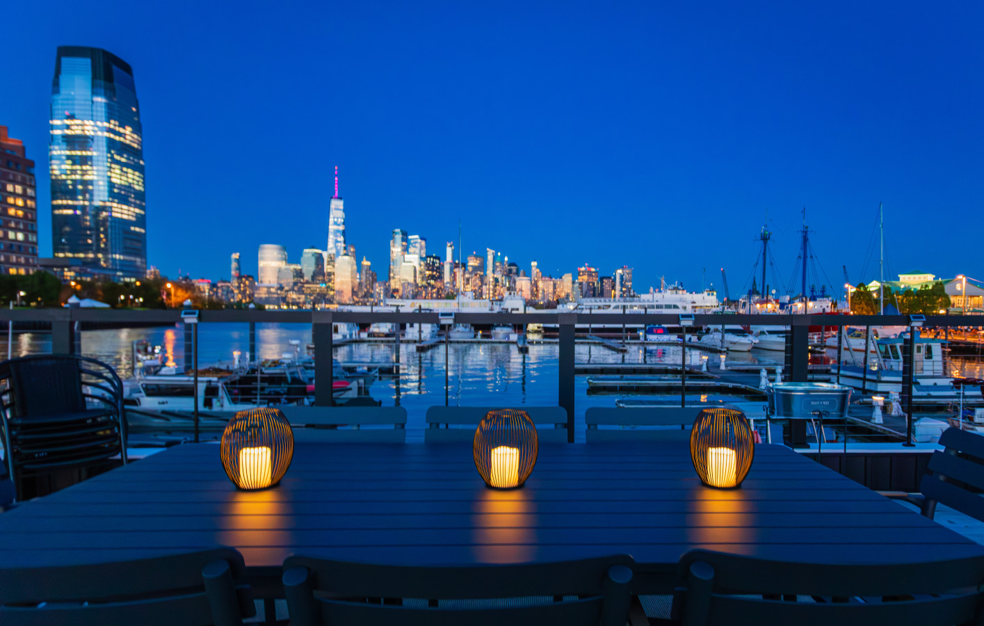 Rooftop dining table at night. Three candlelit lanterns, One World Trade centered on the horizon, the full Manhattan skyline reflected across the Hudson
