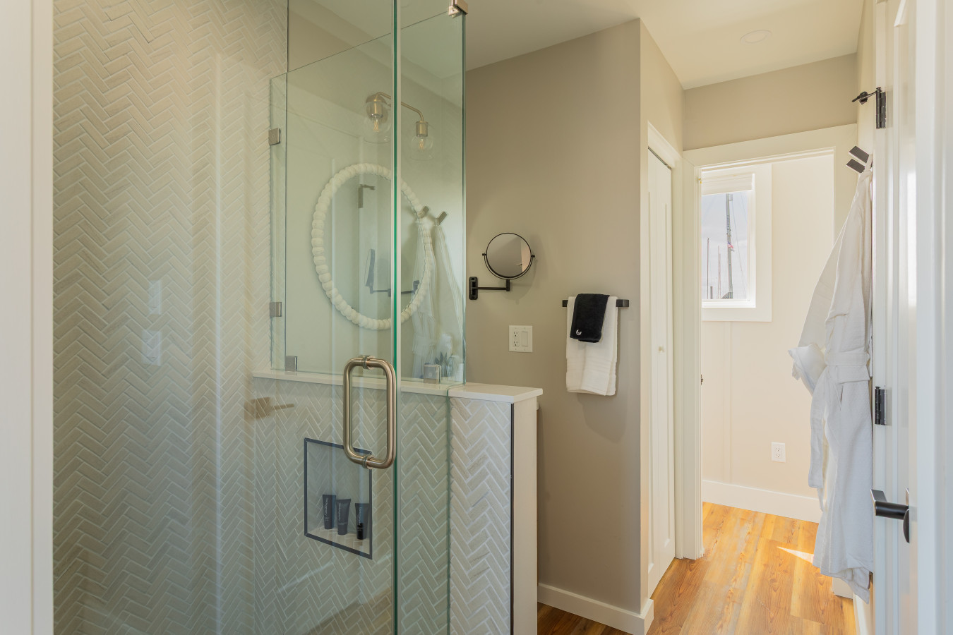 The primary bath features a floor-to-ceiling herringbone tile shower with a glass enclosure, complimentary amenities, and FLOHOM robes waiting on the door.