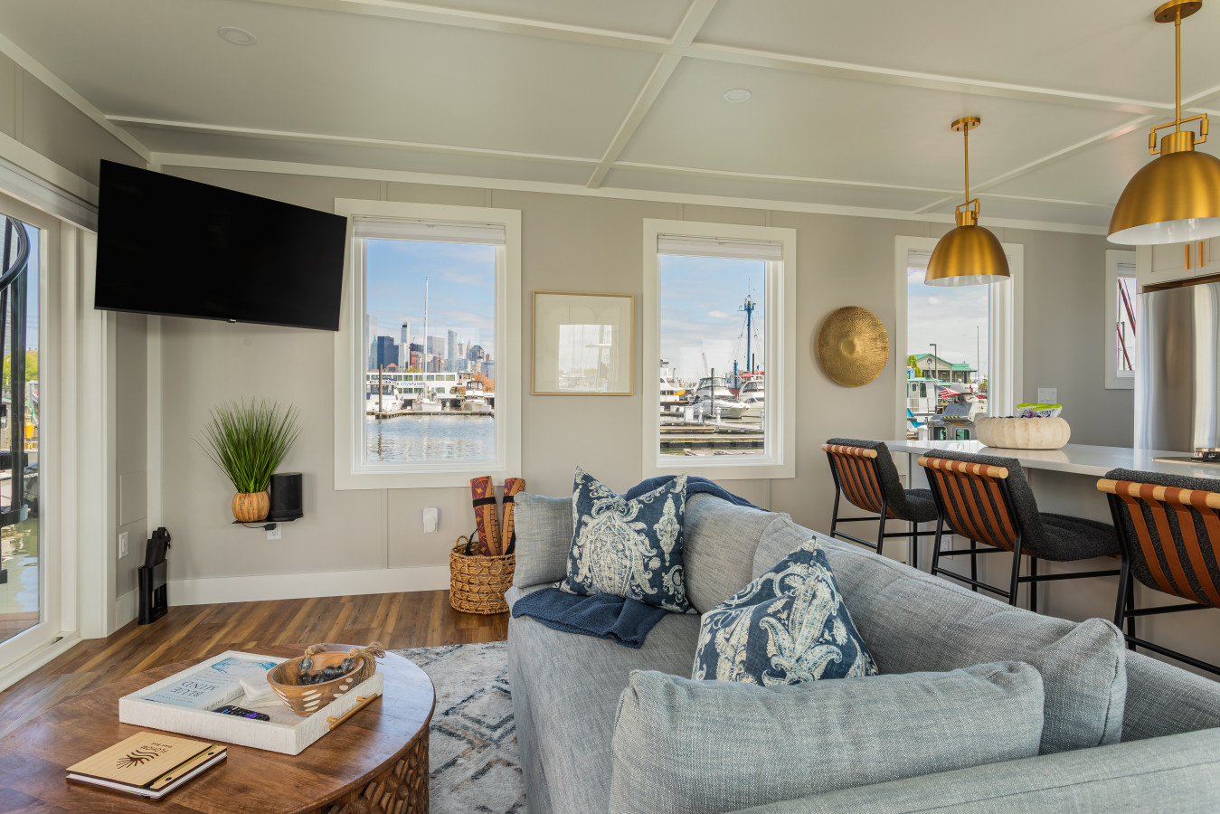 The living area looks out directly at the Manhattan skyline, with a wall-mounted TV, bar seating, and a sliding glass door opening to the water.