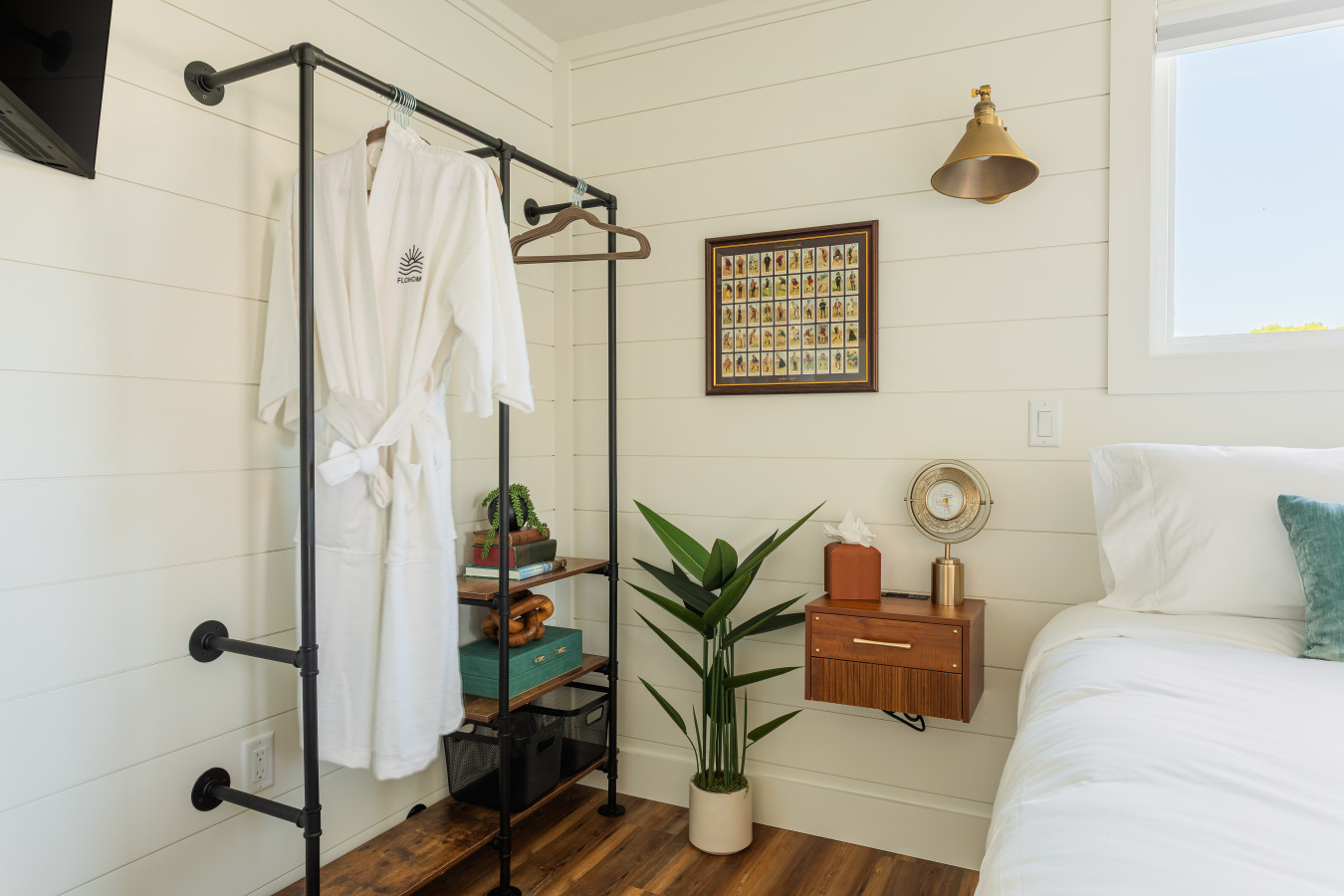 Master suite detail with a FLOHOM kimono robe on the open wardrobe rack, brass sconce lighting, floating nightstand, and shiplap walls dressed with vintage coastal art.