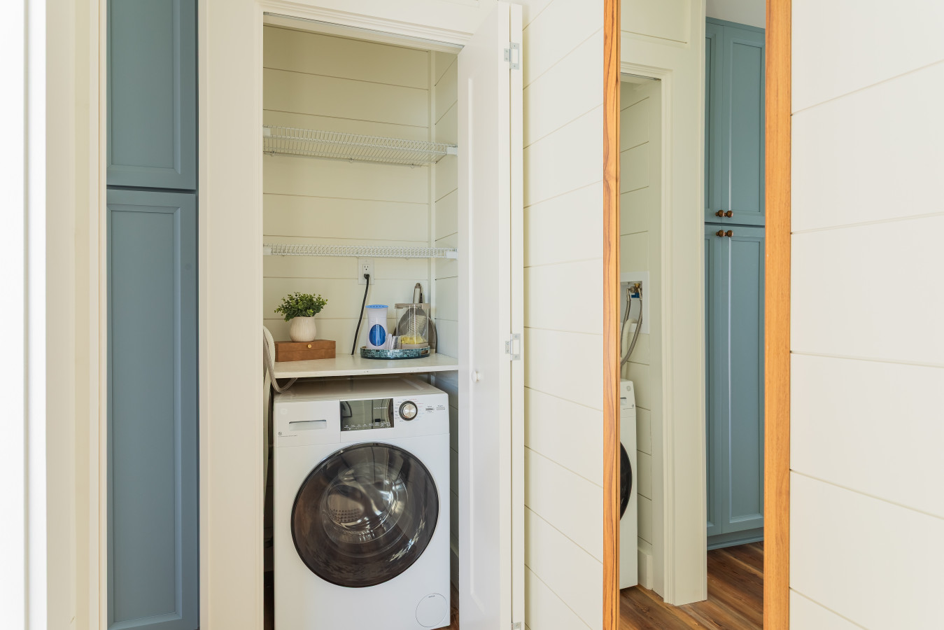 An in-unit washer and dryer closet tucked between the blue-gray cabinetry panels in the hallway, with shelving above and the passage to the bedrooms just beyond.