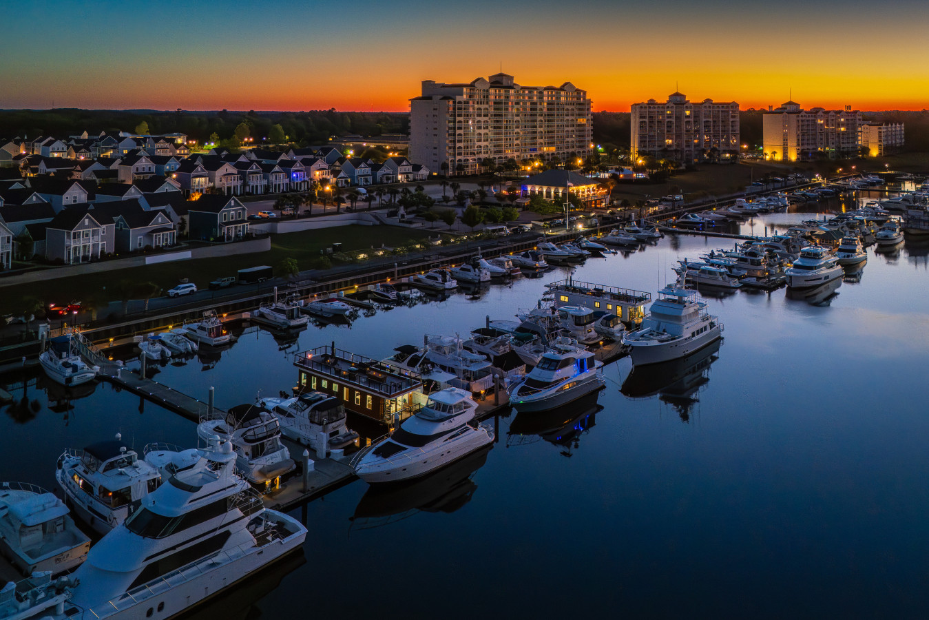 An aerial view of Barefoot Landing Marina at blue hour, with Selah Sailor docked among the yachts and the Barefoot Resort towers glowing on the horizon.