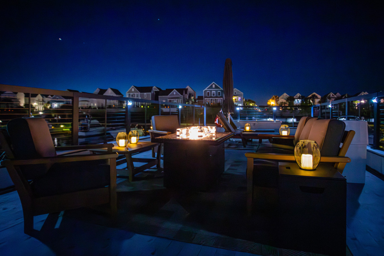 The rooftop fire pit at night, with lounge seating, lantern accents, and the lit waterfront homes of Barefoot Landing across the water.