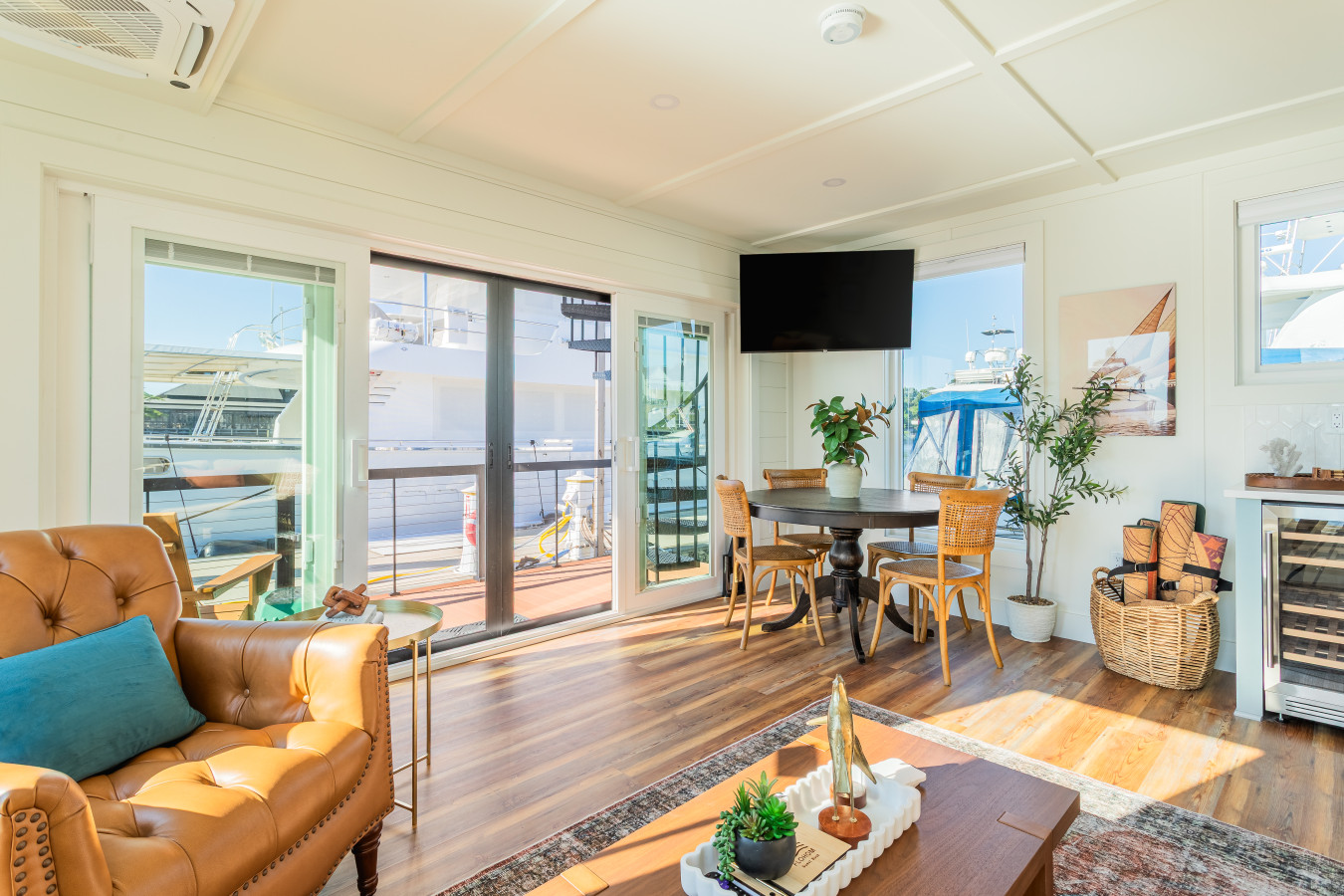 The living and dining area with the tufted leather chair, round dining table, wall-mounted TV, wine fridge, and the entry deck and marina filling the sliding glass doors with natural light.