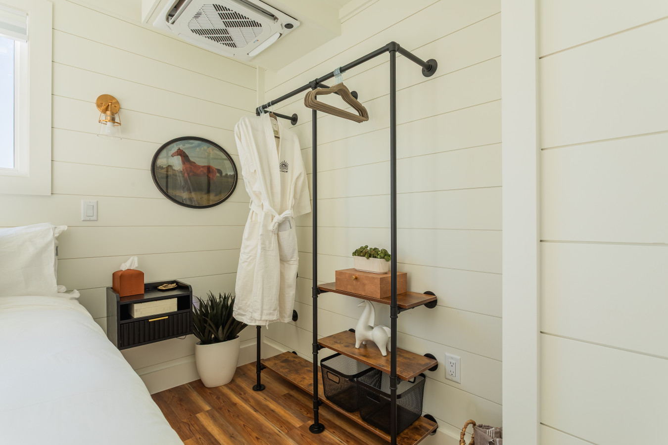 Second master suite detail with a FLOHOM robe on the open wardrobe rack, floating nightstand, brass sconce, and shiplap walls dressed with vintage equestrian art.
