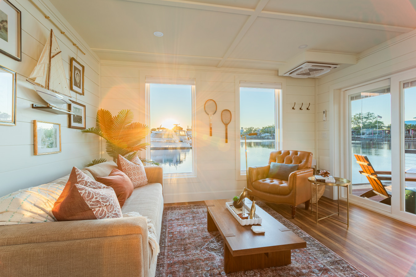The living area at sunrise, with the morning light streaming through marina-view windows across the shiplap walls, vintage maritime gallery, tufted leather chair, and open sliding door to the entry deck.