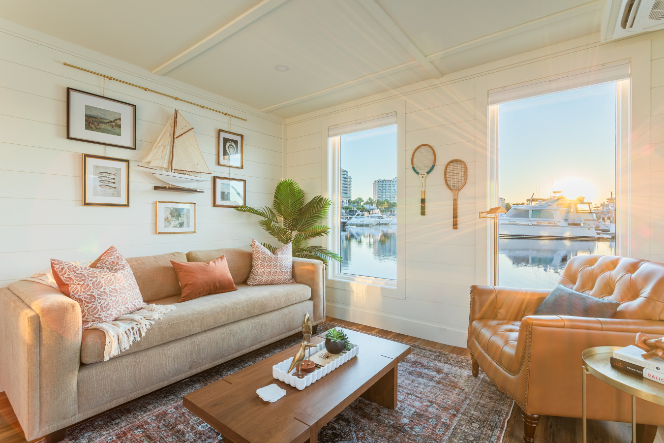 The living area at sunrise, with shiplap walls, a vintage maritime gallery, warm wood floors, and the marina glowing through the windows on both sides of the room.