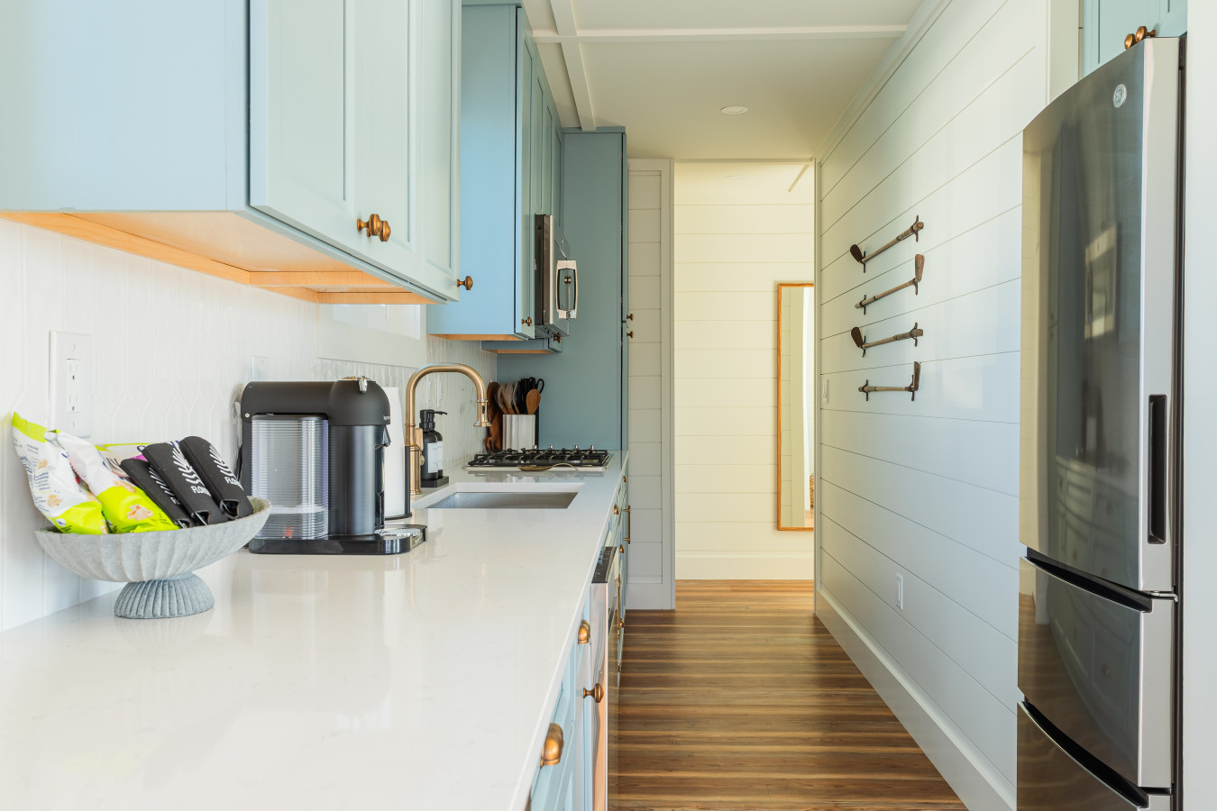 The galley kitchen looking toward the hallway, with quartz counters, a Nespresso maker, brass faucet, gas cooktop, stainless refrigerator, and shiplap walls with nautical hook accents lining the passage to the bedrooms.