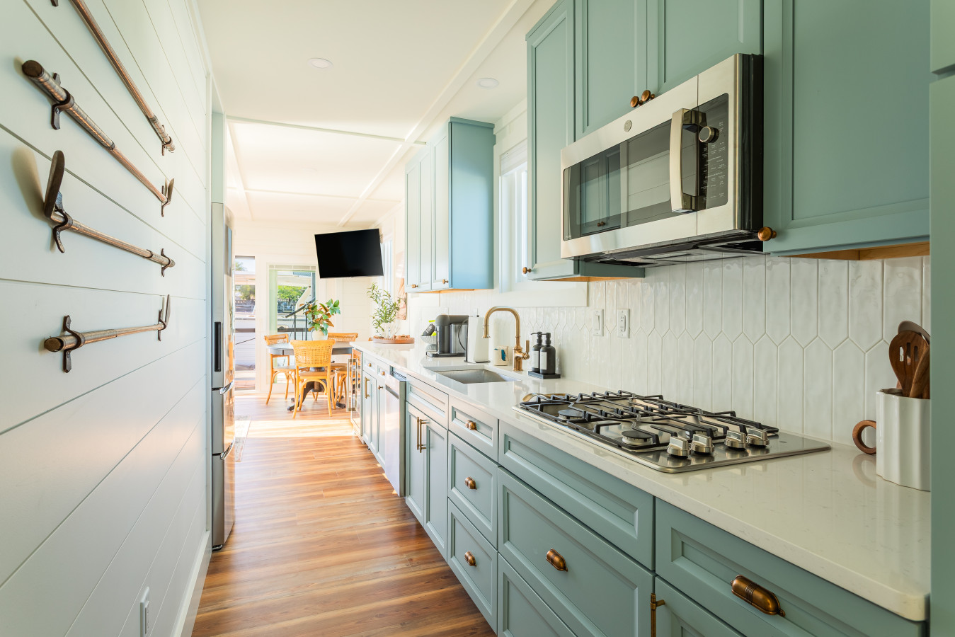 The galley kitchen looking toward the dining area, with blue-gray cabinetry, brass hardware, a gas cooktop, quartz counters, and a white geometric tile backsplash running the full length of the space.