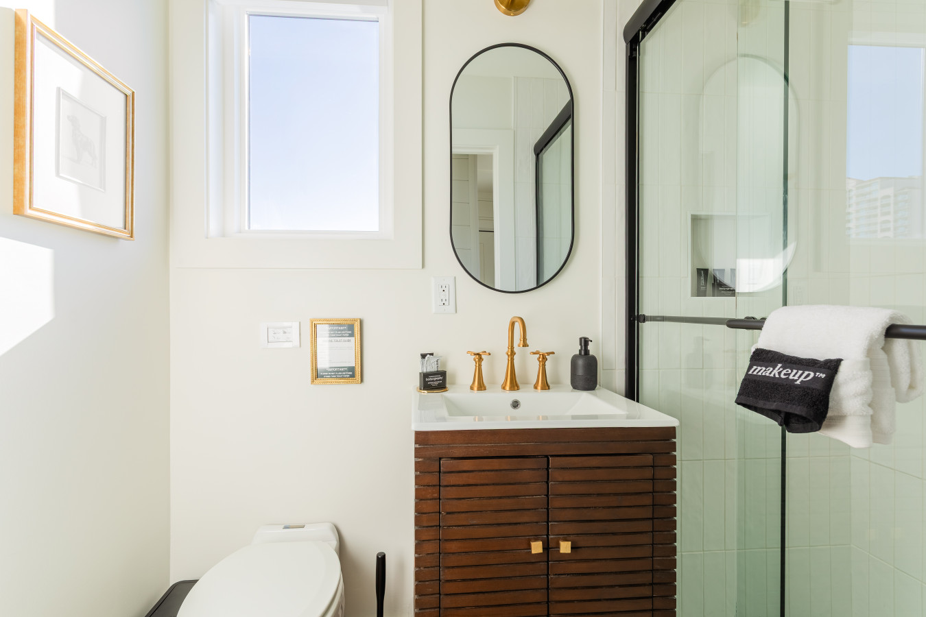 Master bathroom with a walnut vanity, brass fixtures, oval mirror, walk-in glass shower, and a dedicated makeup towel, all finished in soft white tile and shiplap.
