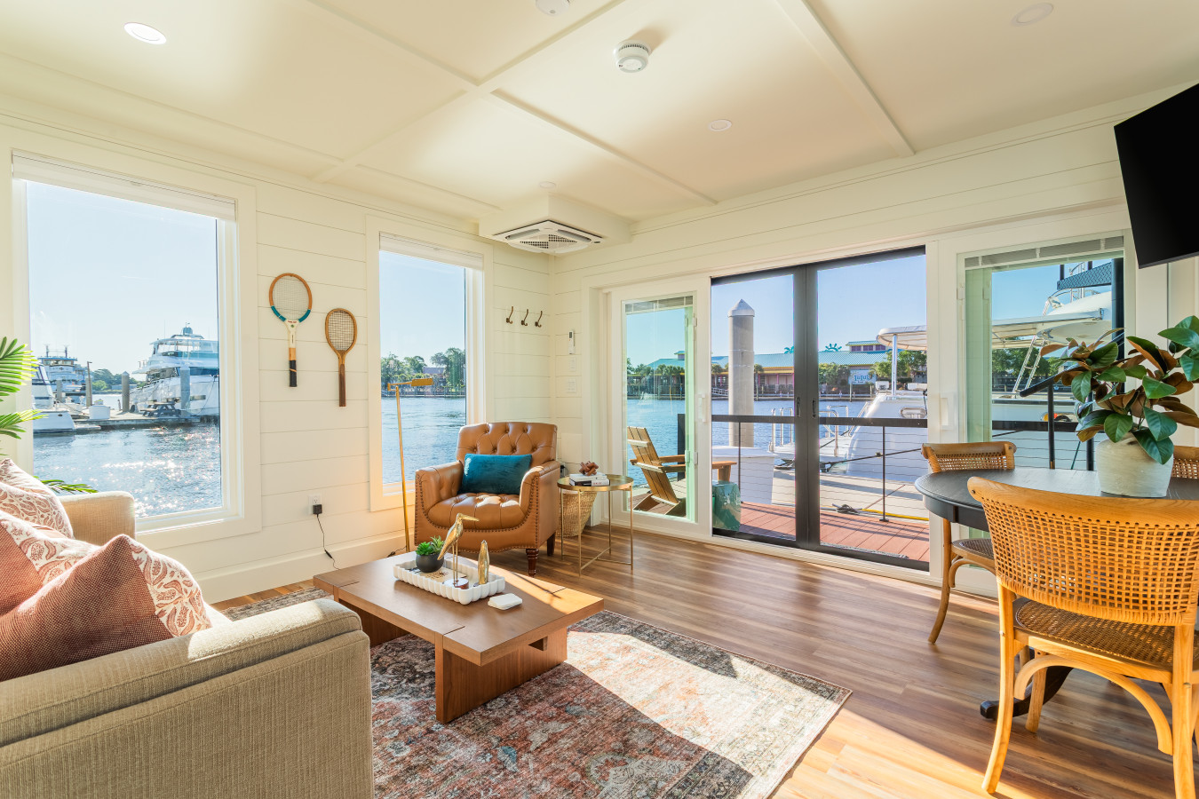 The sun-filled living area with marina views on three sides, shiplap walls, a tufted leather chair, warm wood floors, and the entry deck and waterway visible through the open sliding glass door.