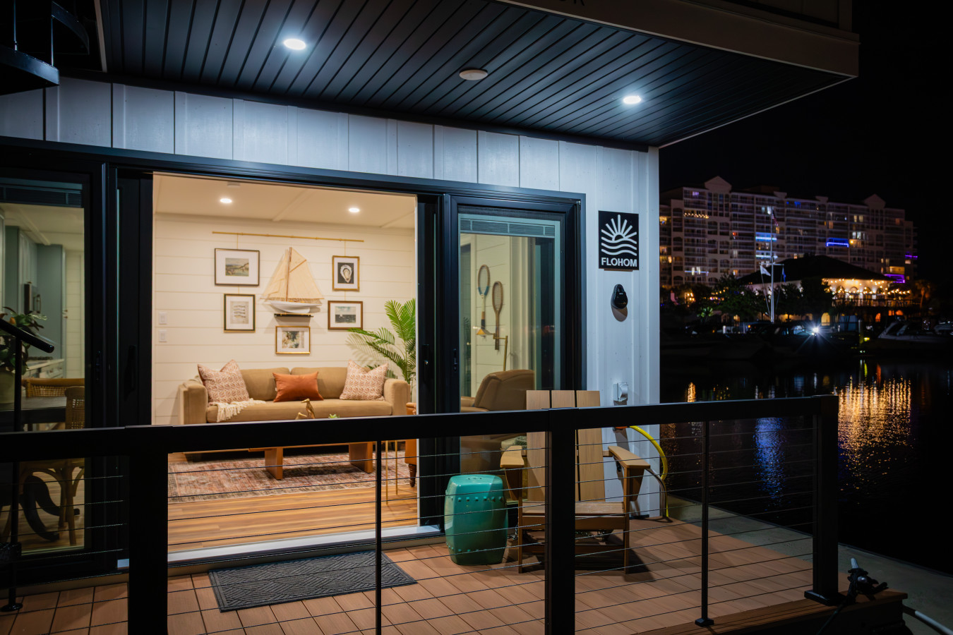 The entry deck at night, with the living area's shiplap walls, maritime gallery, and coastal furnishings glowing through the open sliding glass doors and the Barefoot Resort lit across the waterway.