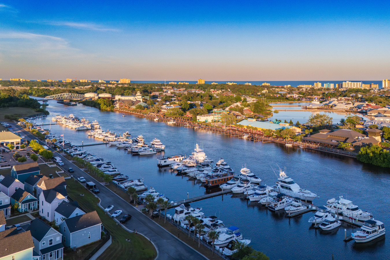 An aerial view of Barefoot Landing Marina at golden hour, with the Barefoot Landing entertainment district directly across the Intracoastal and the Atlantic coastline stretching along the horizon.