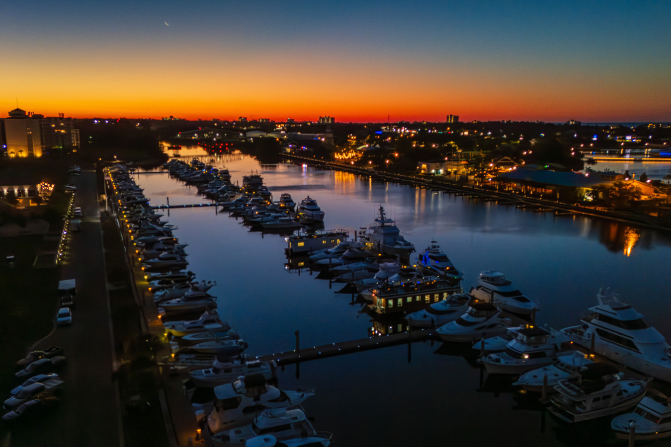 An aerial view of Barefoot Marina at blue hour before sunrise, with the Intracoastal Waterway reflecting the city lights and a crescent moon still visible in the pre-dawn sky.