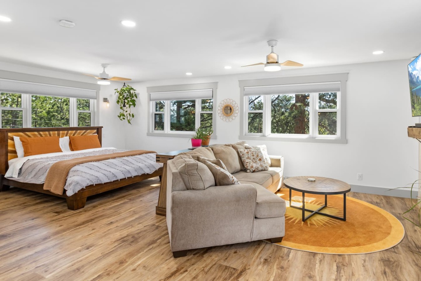 Open interior with a wooden bed frame and orange bedding beside a beige sofa facing a round coffee table on an orange rug. Re