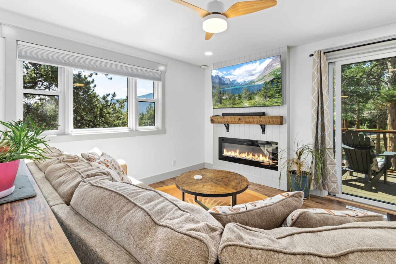 Beige sectional sofa facing a round wooden table, a wall-mounted television displaying a mountain landscape, and a lit glass