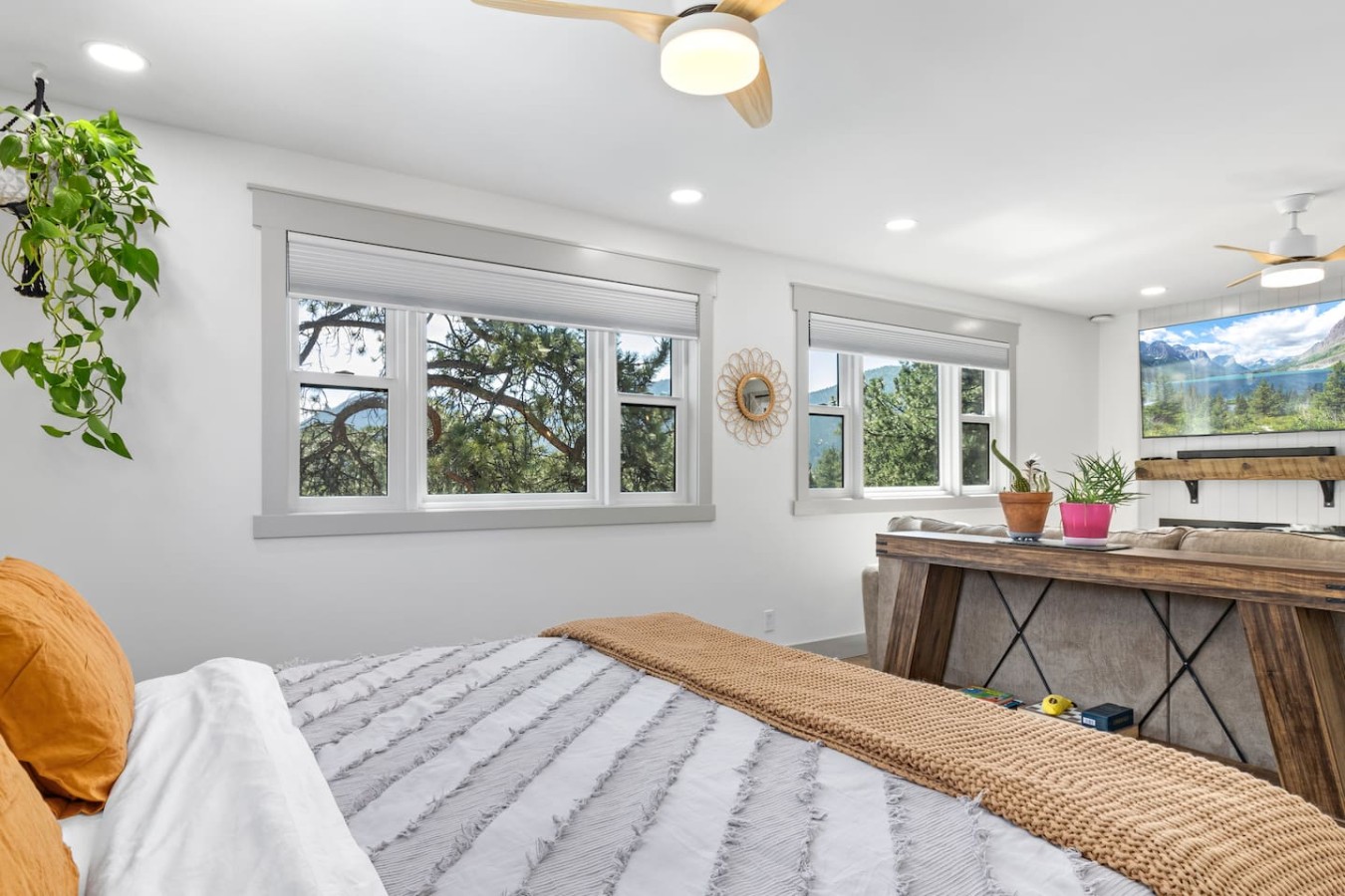 A bed with white and gray bedding and a tan woven throw faces a rustic wooden counter. Two windows, potted and hanging plants