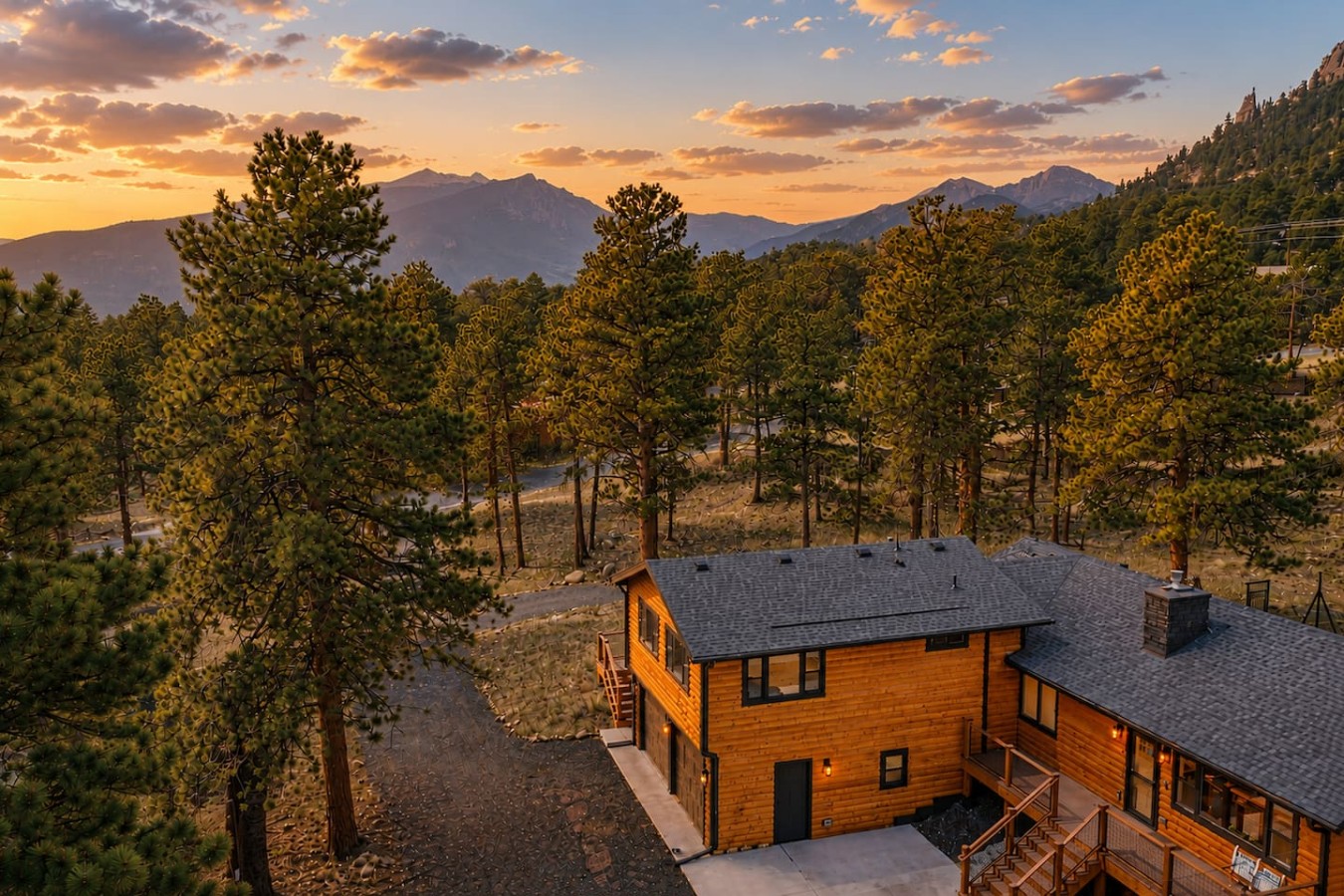 A wood-sided building with dark roofs and exterior stairs stands among tall trees, with layered mountains and orange-lit gray