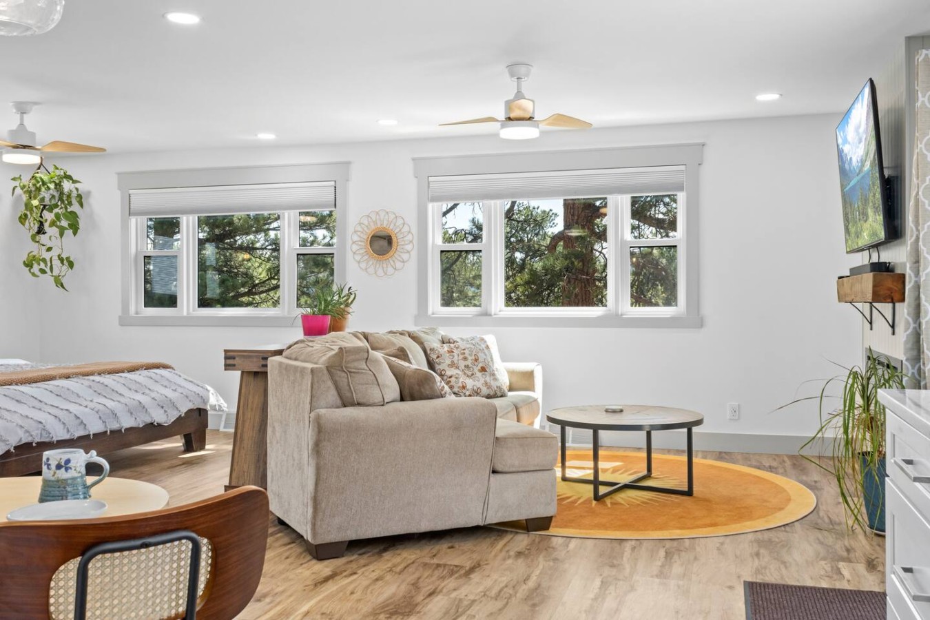 A beige sofa faces a wall-mounted television beside three windows. A round rug and coffee table sit on the wood-look floor, a