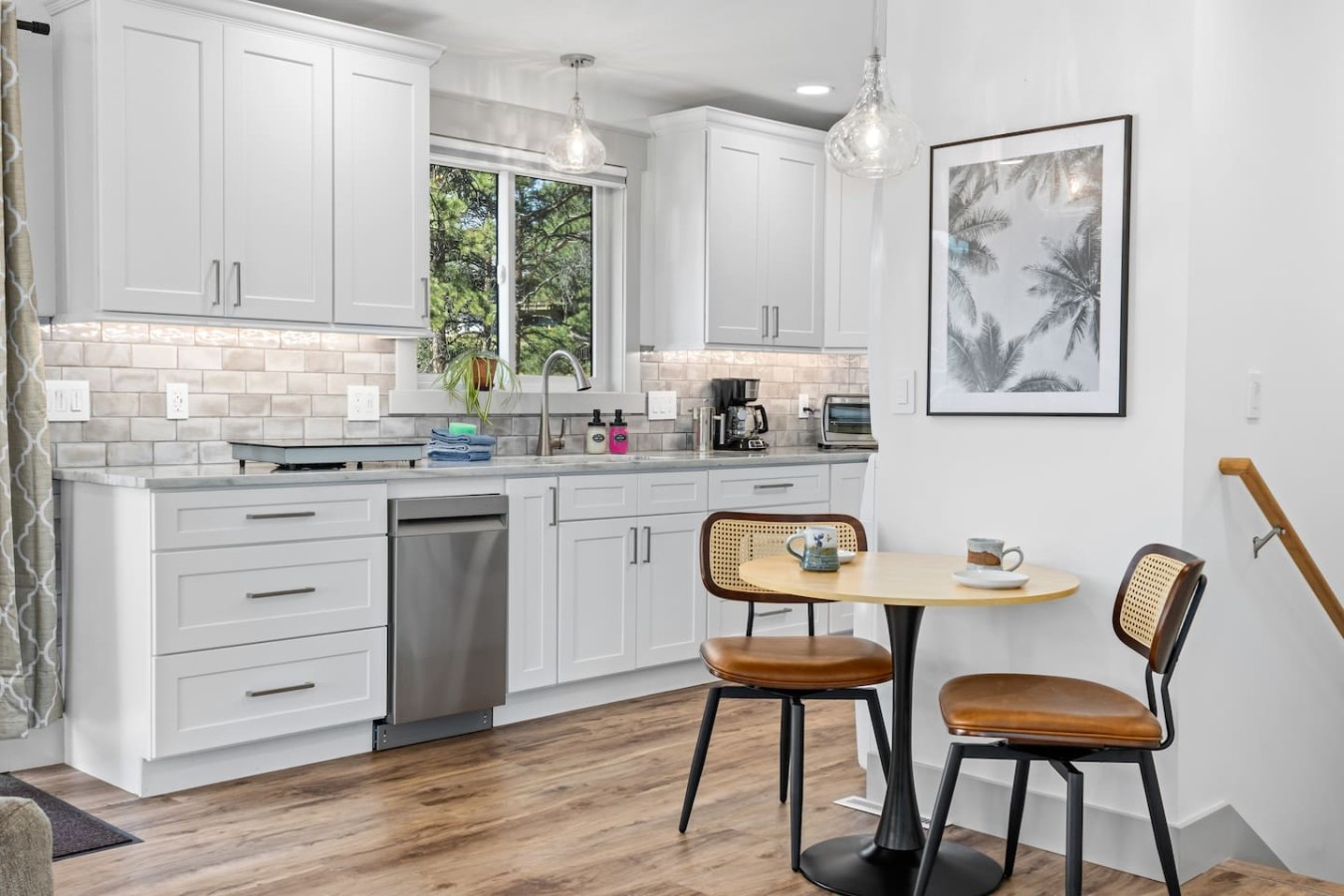 White cabinets line the wall beneath a window, with a sink and stainless-steel dishwasher below. A round table with two brown