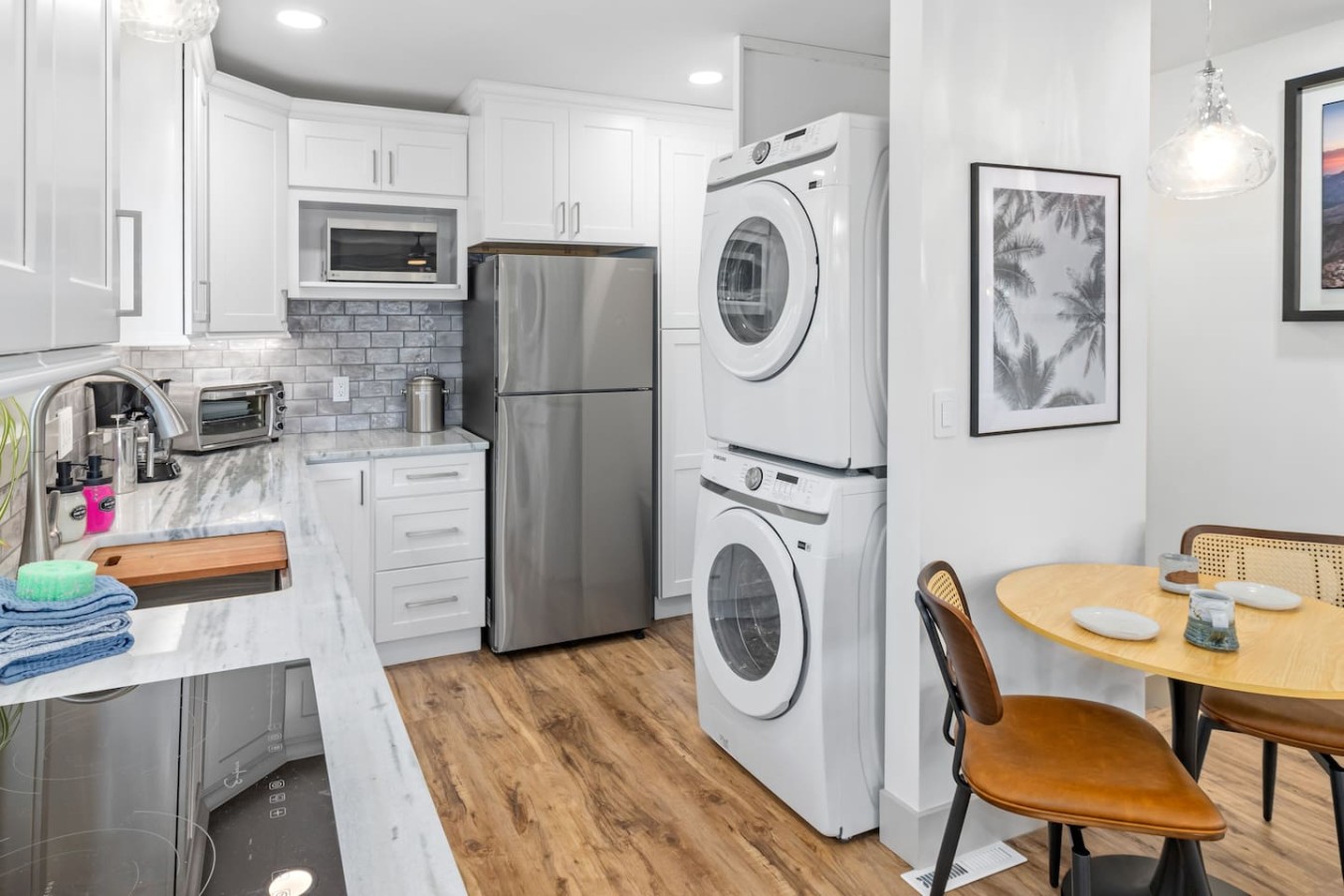 White cabinets and counters beside a refrigerator and stacked front-loading machines, with a round wooden table and chairs on