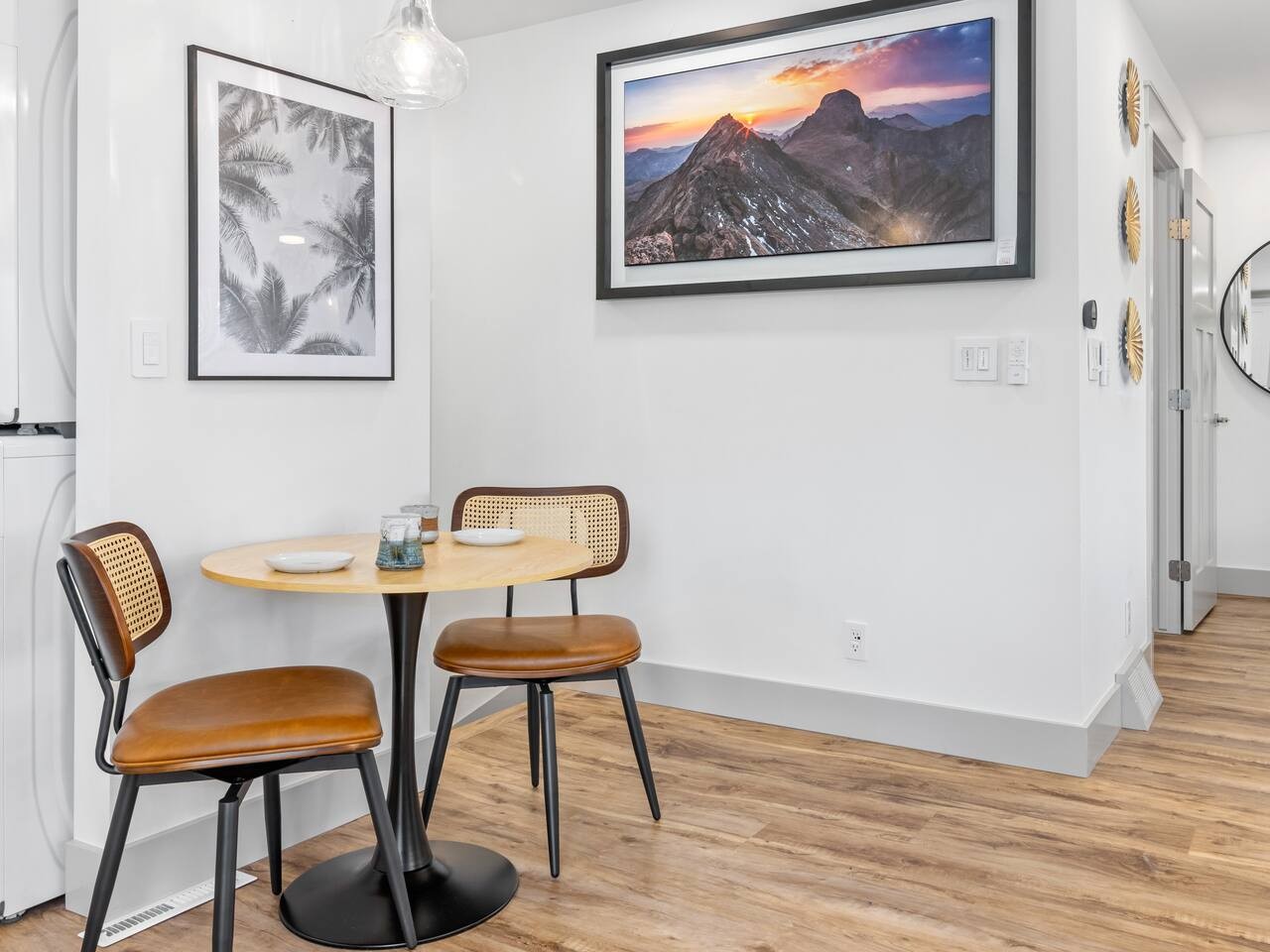 Two brown chairs with woven cane backs flank a small round wooden table holding a bowl and small items. Framed palm artwork