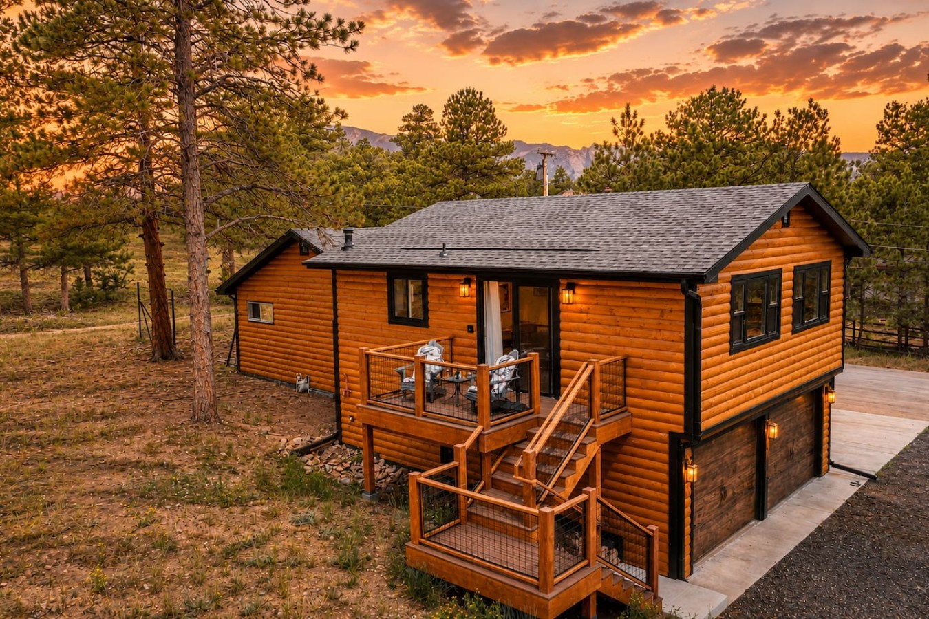 A wood-sided building with a dark shingled roof, exterior lights, raised deck, railings, and stairs among pine trees.