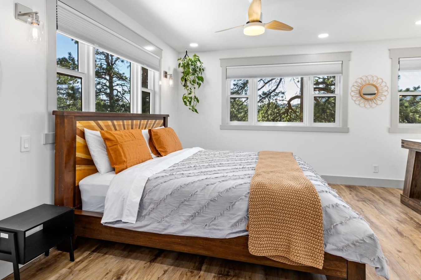 Wooden bed beside several windows, with gray and white bedding, orange pillows, a textured orange throw, a black side table