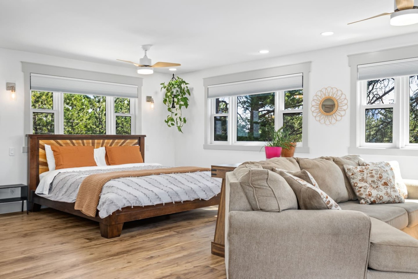 Bright interior showing a wooden bed with orange and white pillows and layered bedding beside a gray sectional sofa. Multiple