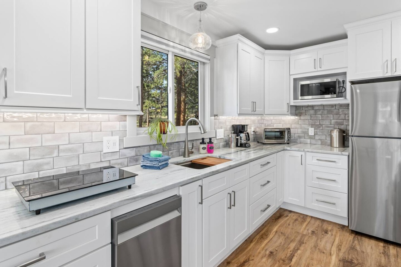 White cabinets surround a sink beneath a window, with a dishwasher, refrigerator, microwave, toaster oven, and gray subway-t件