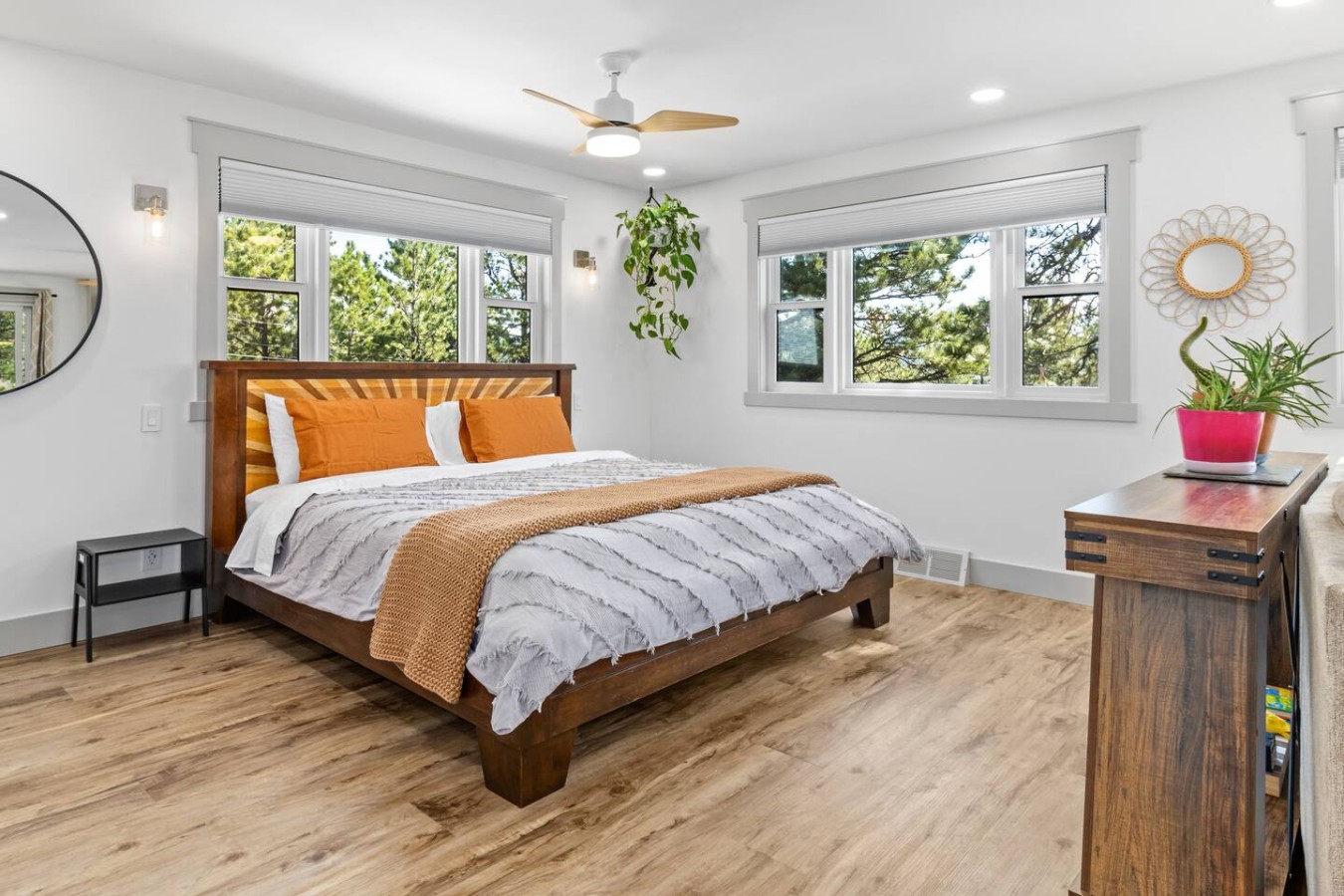 Wooden bed with orange pillows and gray bedding between two windows, with a ceiling fan overhead, plants, mirrors, and a dark