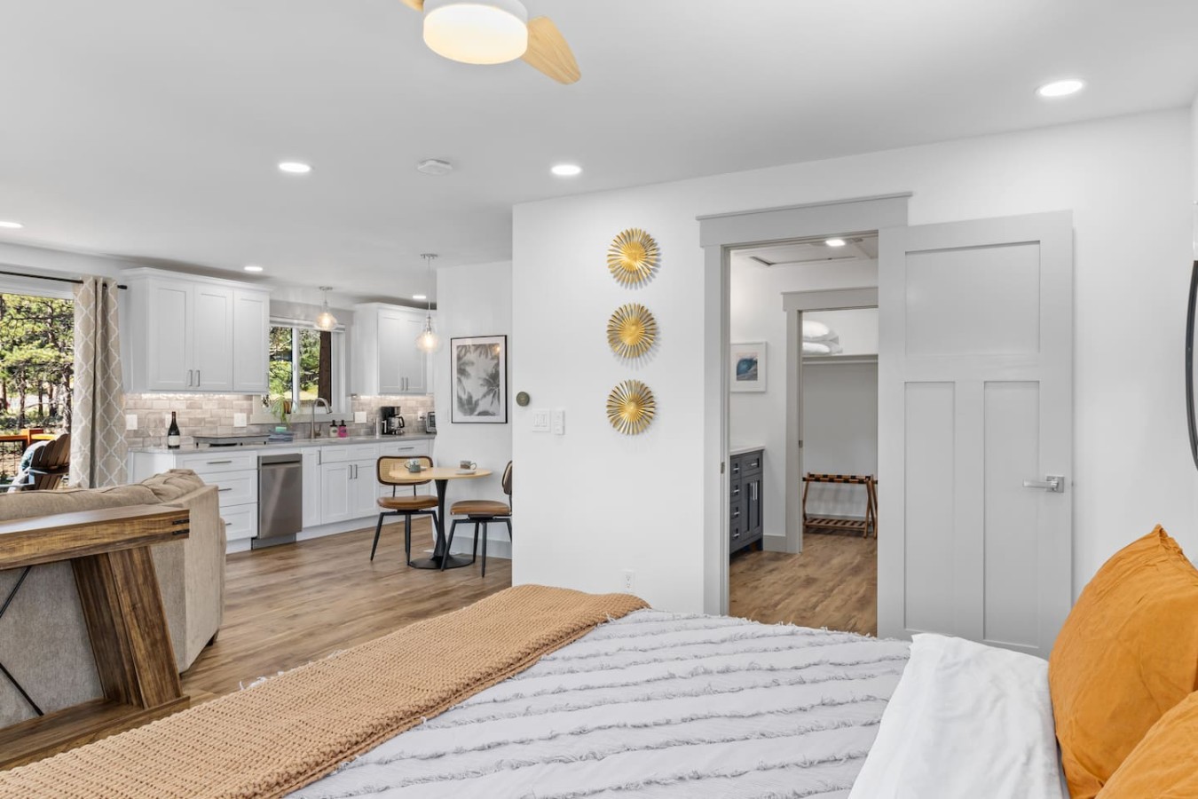 Bed with white and orange bedding beside a gray door, with a table, chairs, white cabinets, and appliances beyond.
