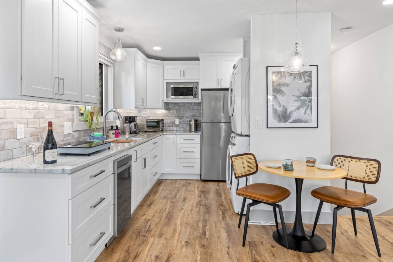 White cabinets and counters line the left wall beside stainless steel appliances. A round table with two cane-back chairs, a_