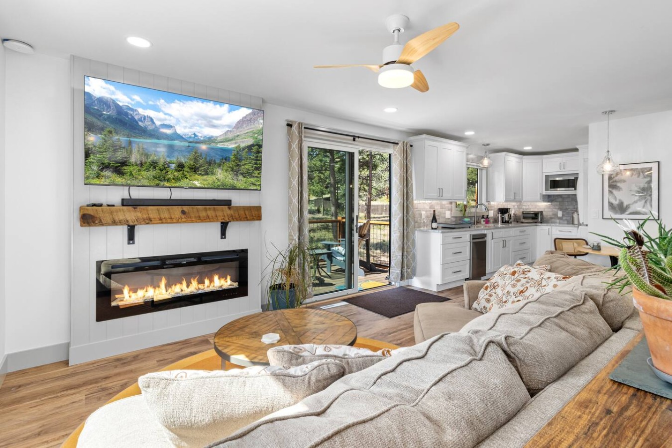 Gray cushioned seating faces a black linear fireplace beneath a wood mantel and wall-mounted screen displaying mountains. A p